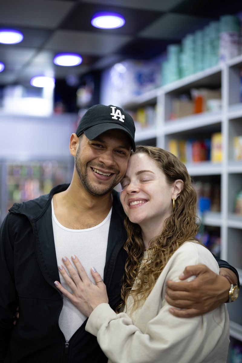 A man in an LA cap holds a smiling woman in a cream sweater close inside a South Street shop, her ring hand on his chest.