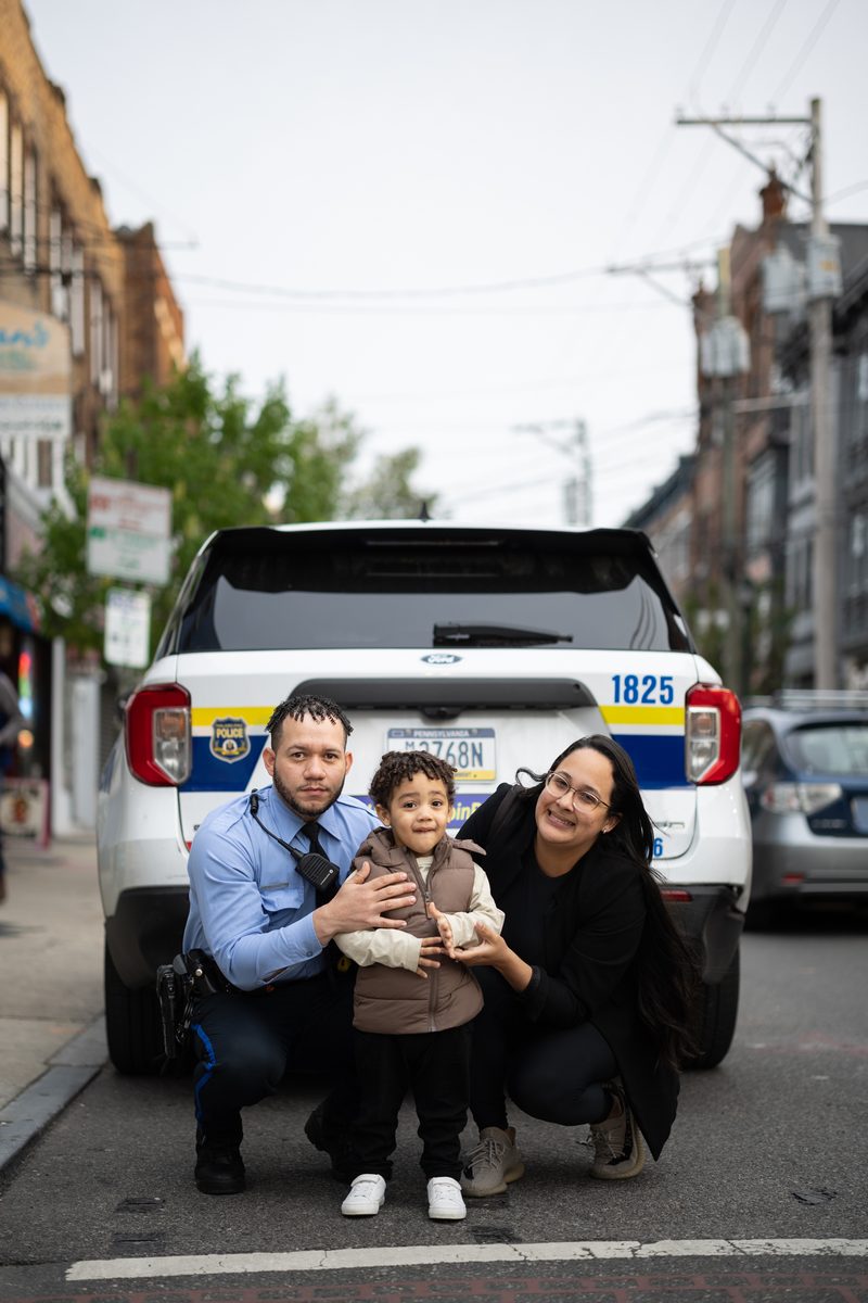 A Philadelphia police officer crouches with his young son and partner in front of a police SUV on South Street.