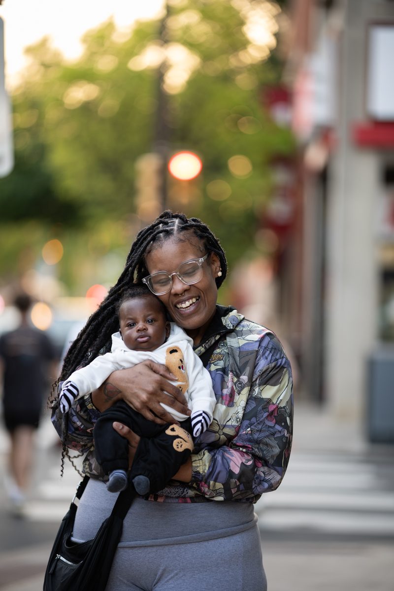 A woman with braids and glasses cradles her baby, dressed in white, on a South Street sidewalk at golden hour.