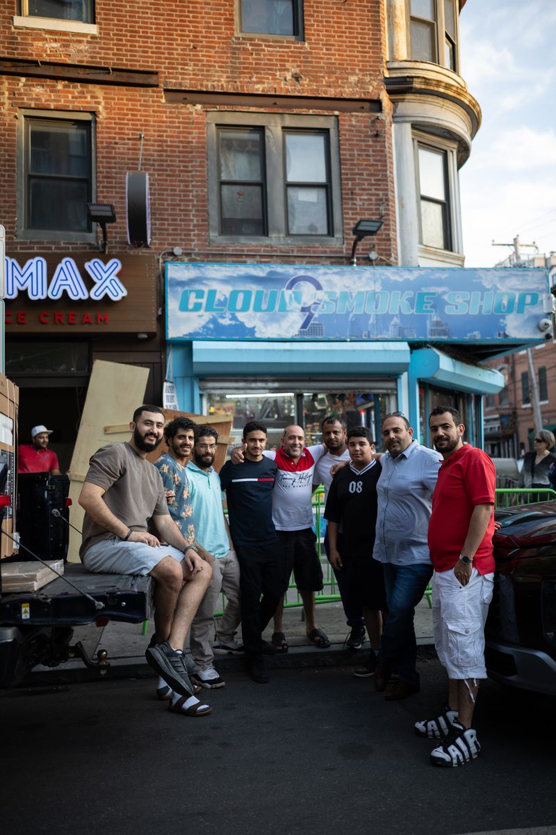 Nine men stand together outside a smoke shop and an ice cream shop storefront on South Street, one seated in a pickup truck tailgate on the left.