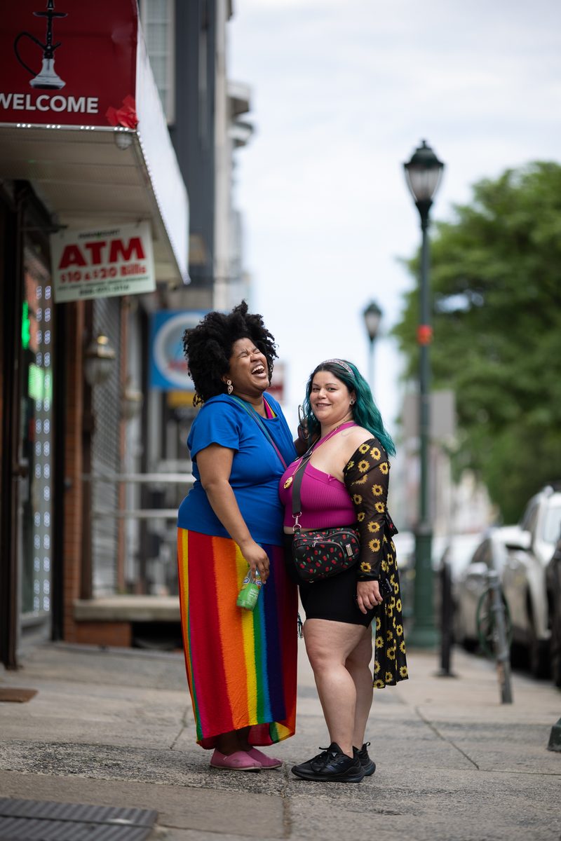 Two friends laugh on a South Street corner, one in a rainbow skirt, the other in a sunflower cardigan with teal hair.