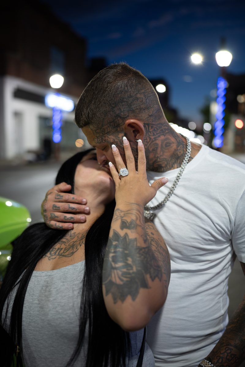 A tattooed man kisses a woman with long black hair, her engagement ring catching the light, on South Street at dusk.