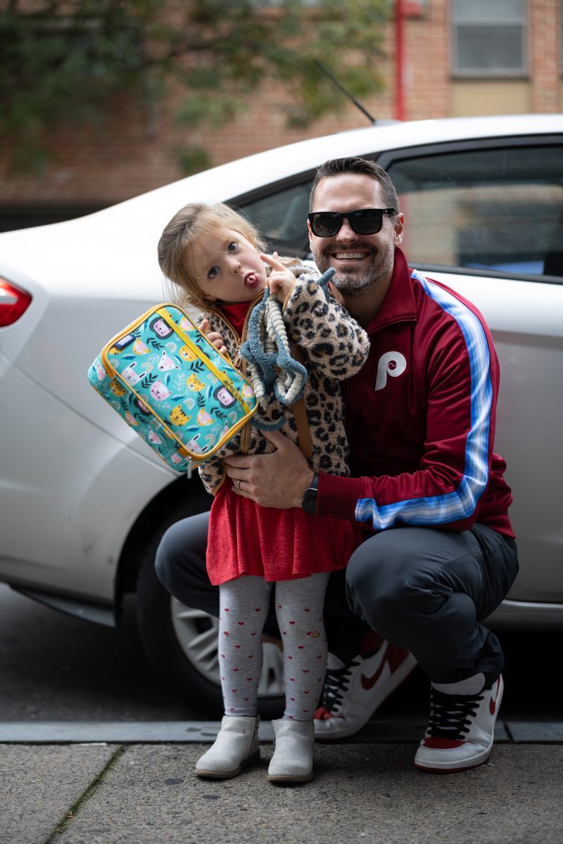 A father in a Phillies jacket crouches beside his young daughter in a red dress and leopard jacket as she makes a funny face.