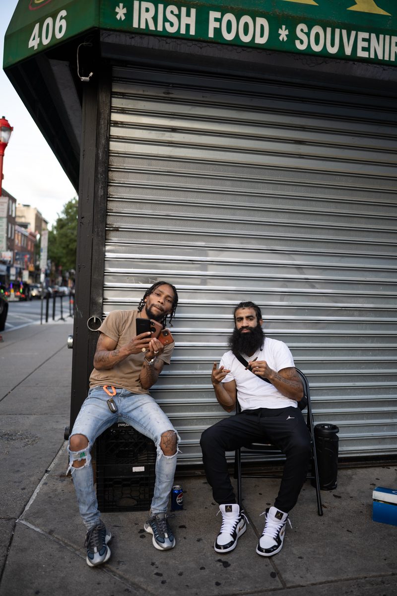 Two men pose while seated on a crate and chair in front of a shuttered South Street Market storefront with an "Irish Food, Souvenirs" sign. One is wearing a tan T-shirt, and one is wearing a white T-shirt. The man in the tan T-shirt flashes three phones like they're playing cards in front of his face.