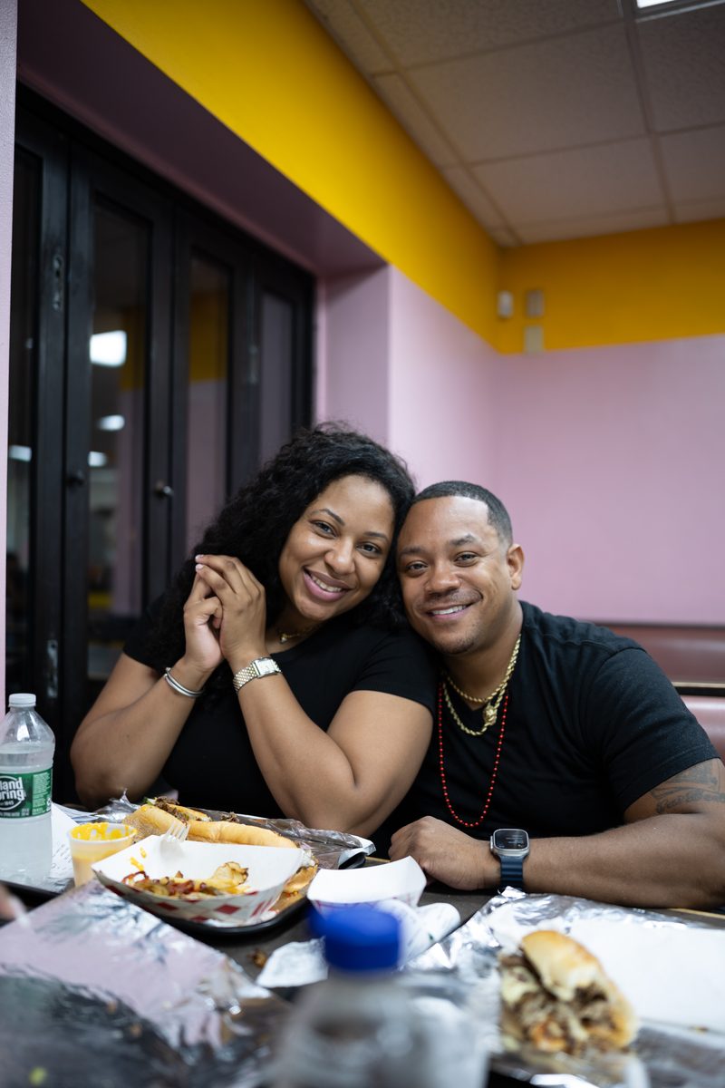 A couple leans cheek to cheek across a table of cheesesteaks and fries at Ishkabibble's on South Street.