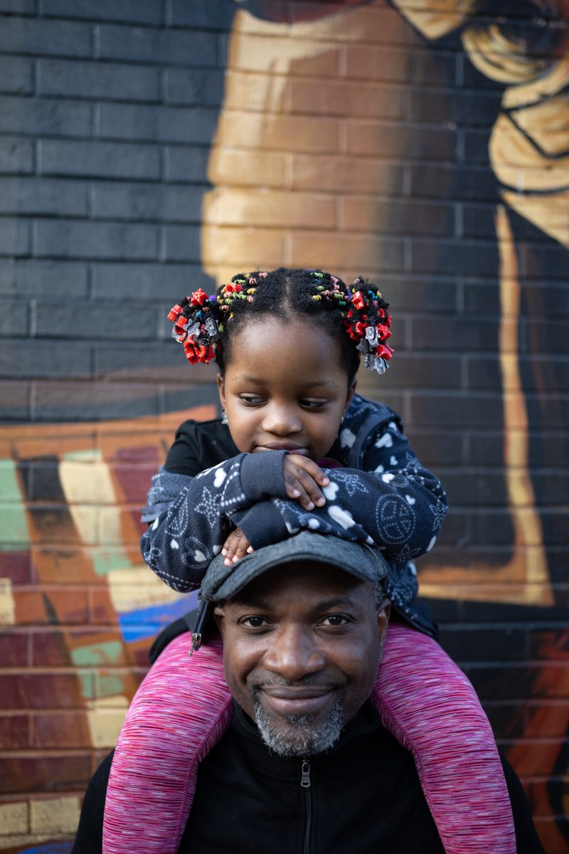 A young girl with beaded braids sits on her father's shoulders in front of the Engine 11 fire station mural on South Street with her hands on his cap.