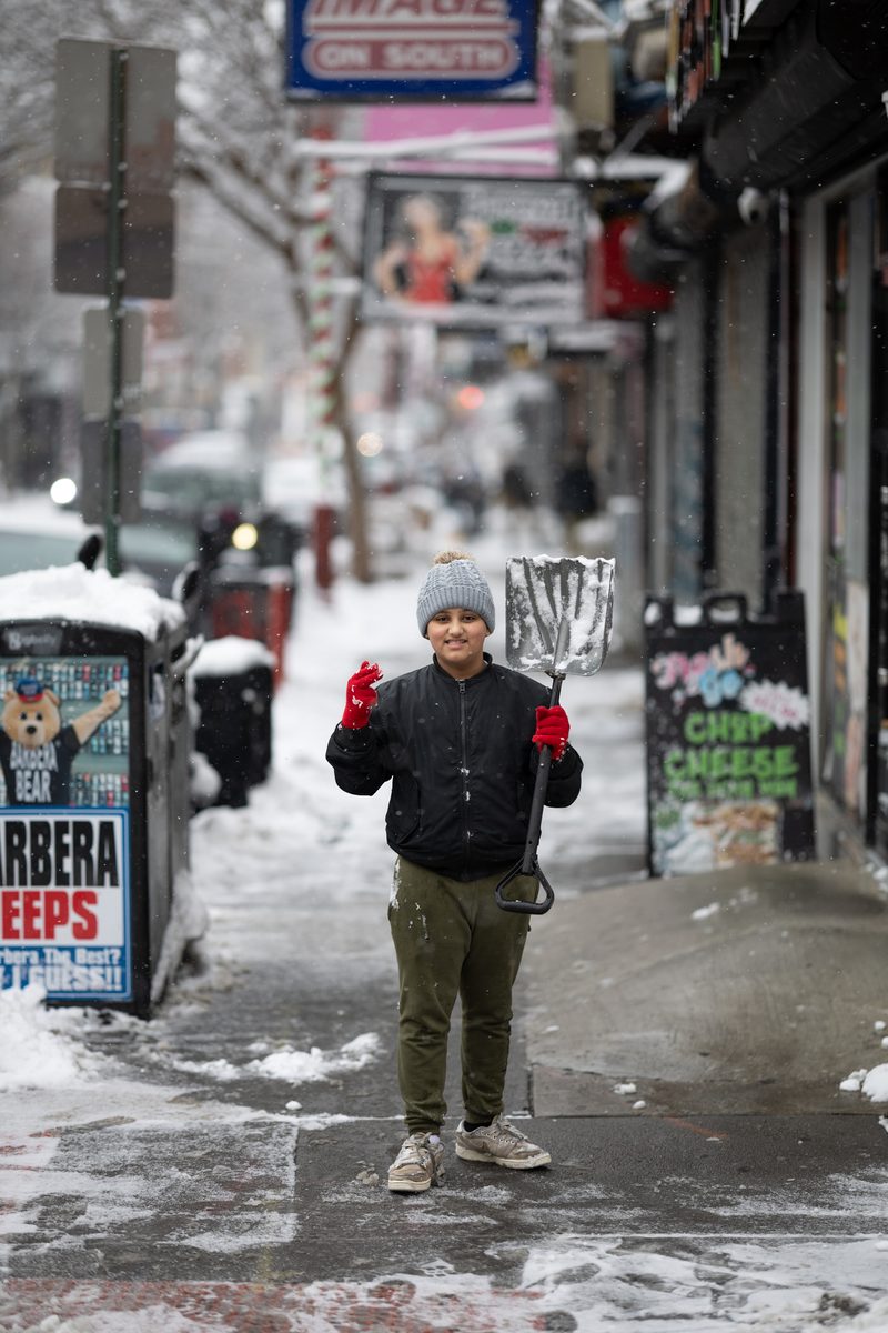 A boy in a gray beanie and red gloves holds a snow shovel on a snow-covered South Street sidewalk.
