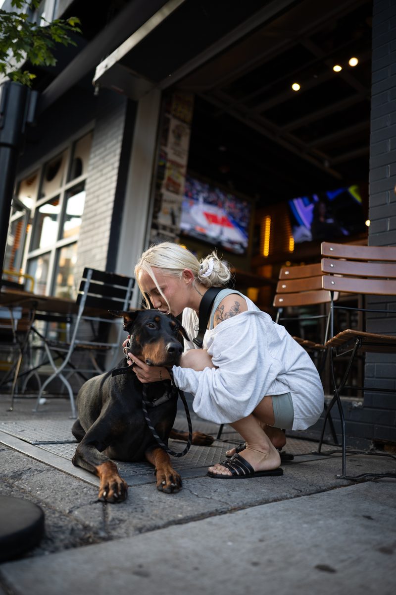 A woman with platinum hair crouches to hug her Doberman on the sidewalk outside of Milkboy bar on South Street.