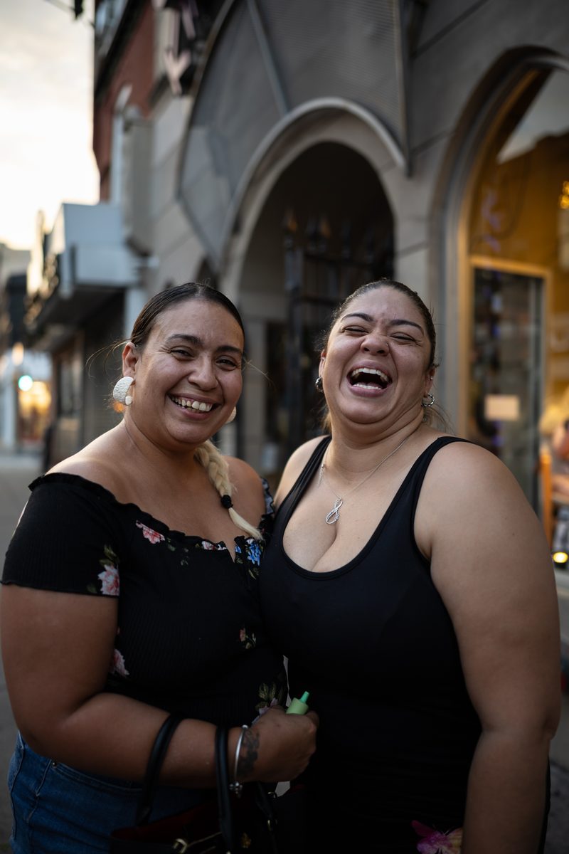 Two women in black tops laugh together on a South Street sidewalk, one in a floral off-the-shoulder top.