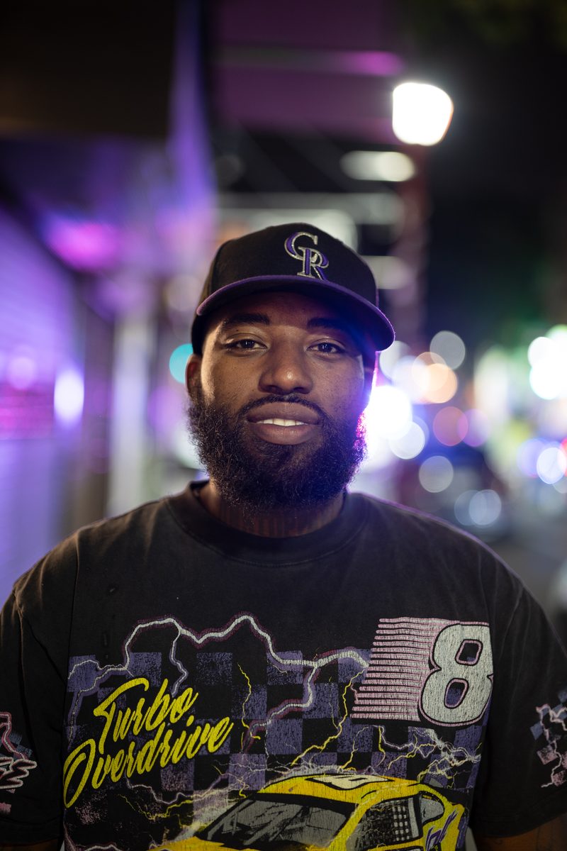 A bearded man in a Colorado Rockies cap and a vintage racing t-shirt smiles under neon lights on South Street at night.