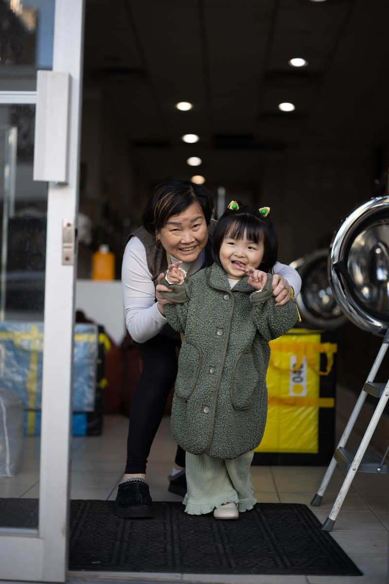 A grandmother hugs a giggling little girl in a green coat and cat-ear headband in a laundromat doorway.