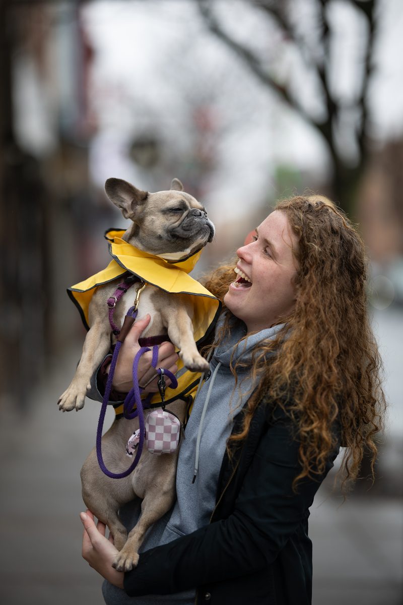 A woman with long curly red hair laughs up at her French bulldog, dressed in a yellow raincoat, held in her arms.