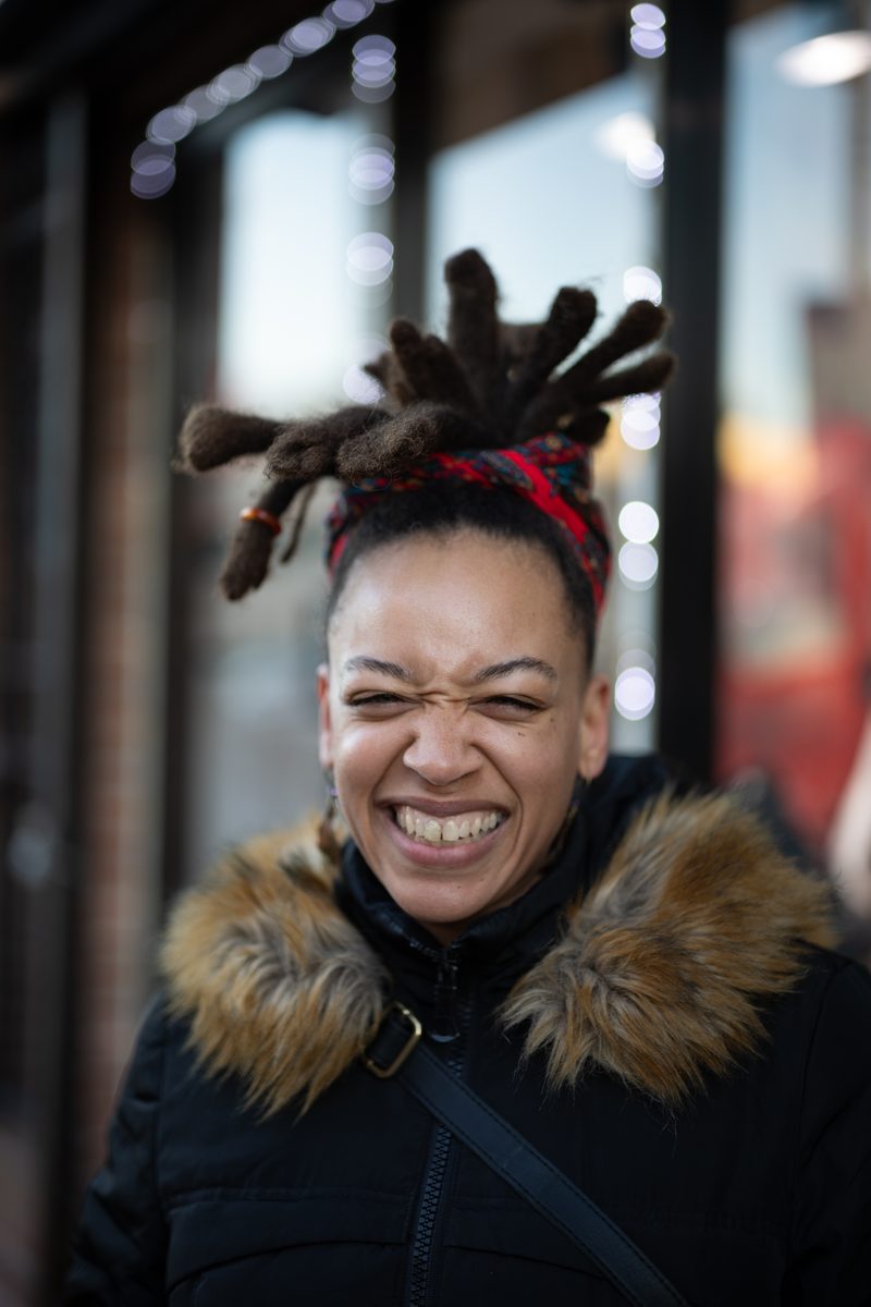 A woman with her locs tied up in a topknot scrunches her face in a big grin, wearing a fur-trimmed winter coat.