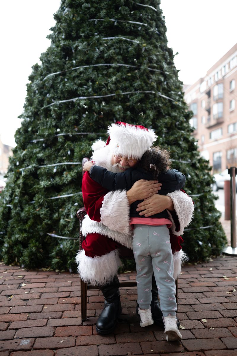 Santa Claus hugs a young girl in front of a tall Christmas tree on a brick plaza.