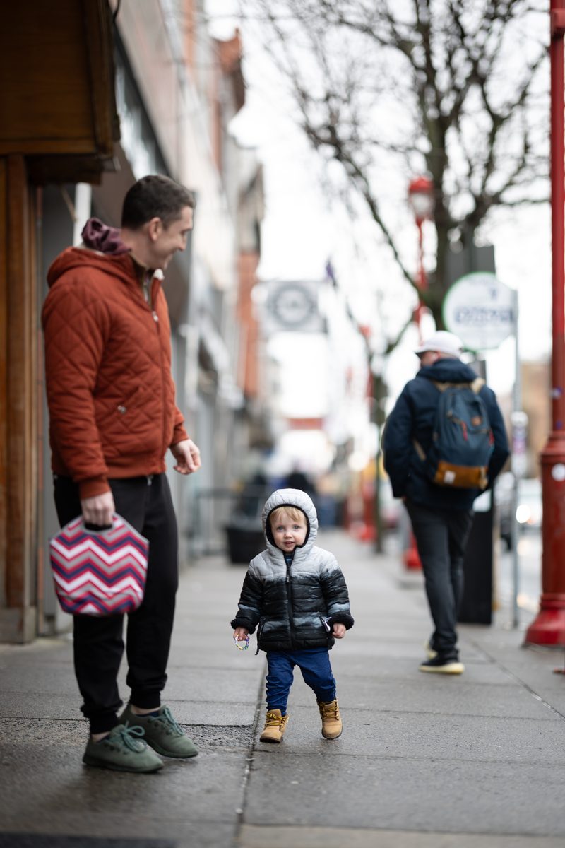 A toddler in a black puffer coat and boots walks towards his father on a South Street sidewalk.