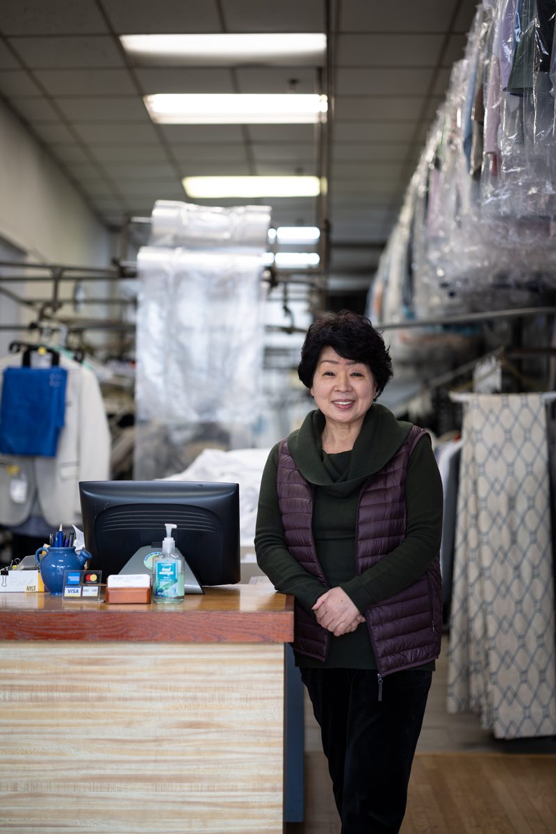 A woman in a purple vest smiles from behind the counter of a dry cleaner, garment bags hanging behind her.