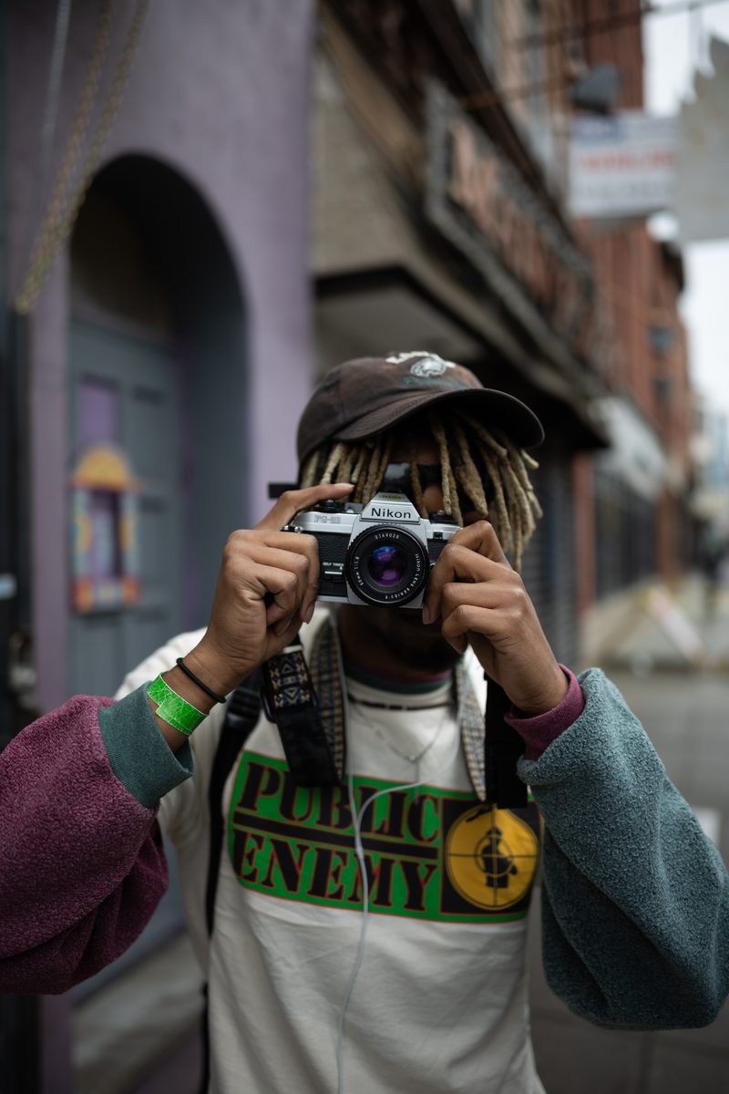 A man in a Public Enemy t-shirt and green wristband holds a Nikon film camera up to his eye on South Street.