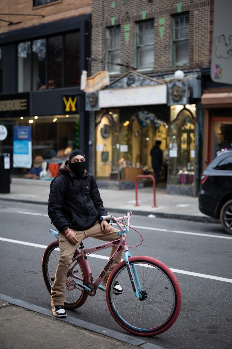 A man in a black balaclava and jacket sits on a pink and blue bicycle in the middle of South Street.