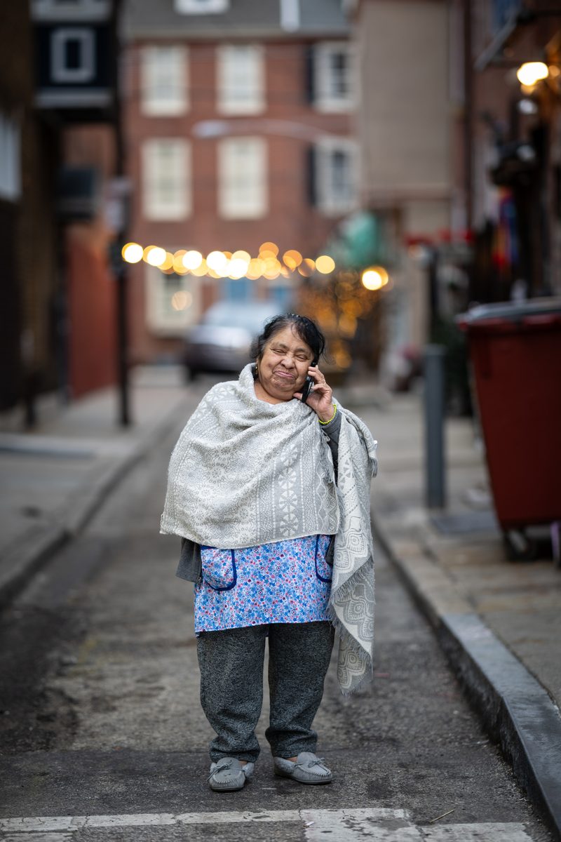 An elderly woman wrapped in a gray shawl talks on her phone while standing in a narrow street strung with lights by Mexitalian Restaurant on South Street.
