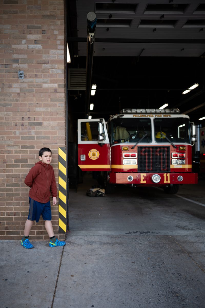 A boy in a red hoodie and blue shorts stands at the open bay of the Engine 11 firehouse on South Street, a fire truck behind him.