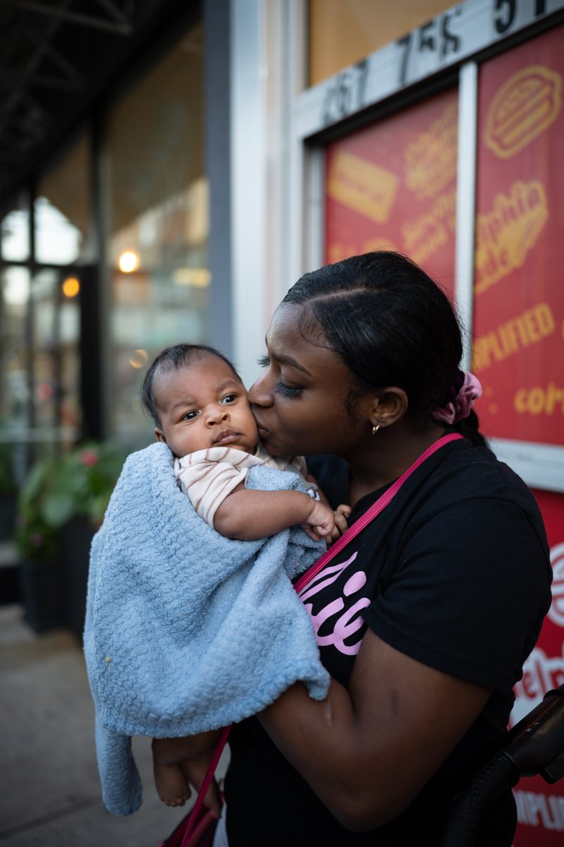 A young mother in a Barbie t-shirt kisses the cheek of her baby, wrapped in a blue blanket, on South Street.