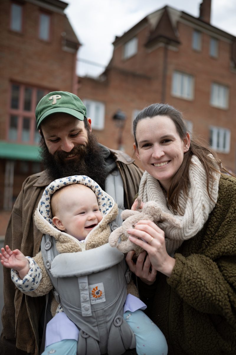 A couple smiles down at their laughing baby in a front carrier, the father in a Phillies cap, on a South Street sidewalk.