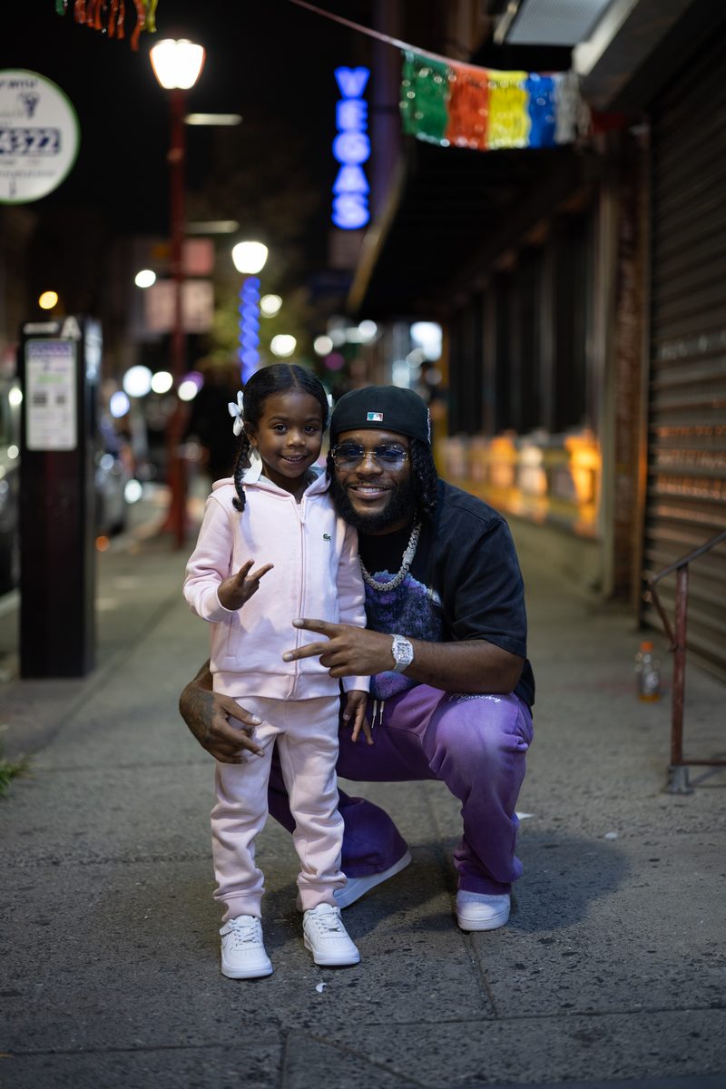 A father in purple pants crouches beside his young daughter in a pink tracksuit, both make peace signs with their hands in a pose on South Street at night.