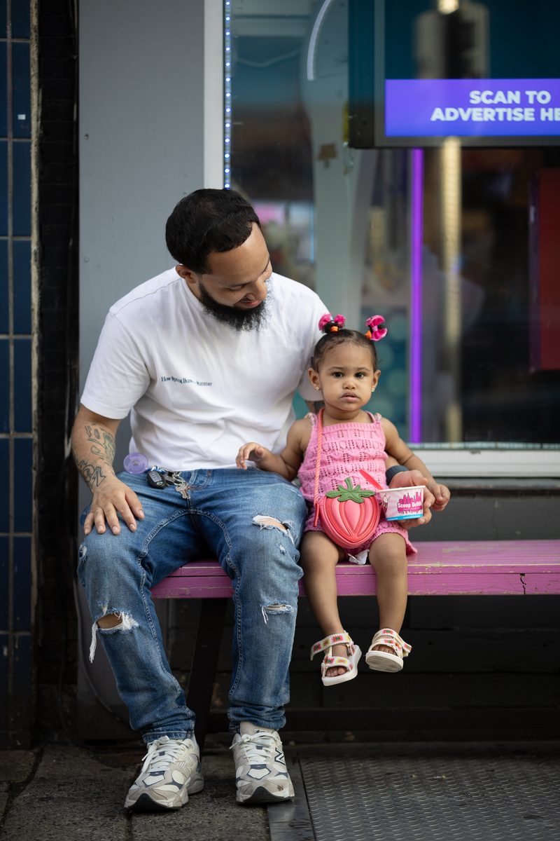A father and his toddler daughter in a strawberry dress sit on a pink bench outside of a South Street ice cream shop called Scoop DeVille.