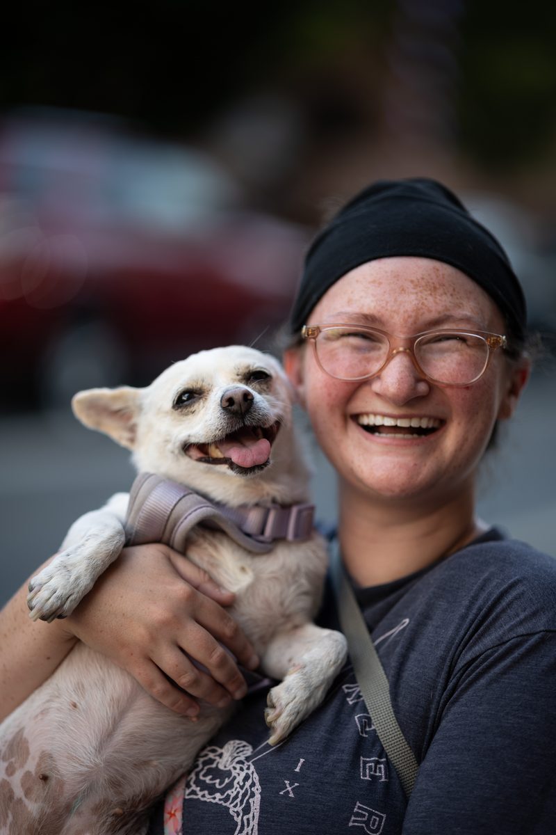 A woman with freckles and glasses laughs while holding her small white dog, who is also smiling, on South Street.