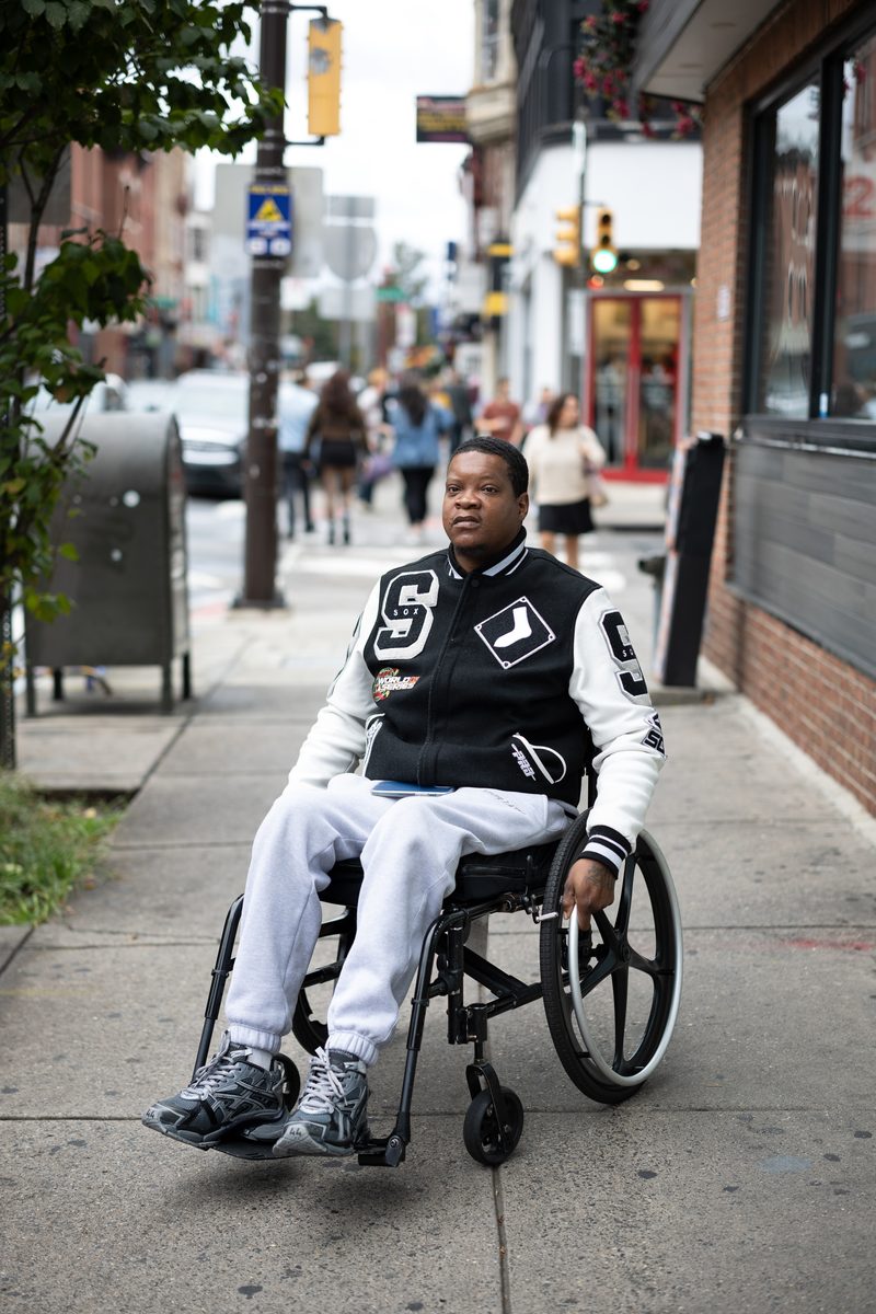 A man in a black-and-white varsity jacket sits in his wheelchair on the South Street sidewalk, traffic lights behind him.