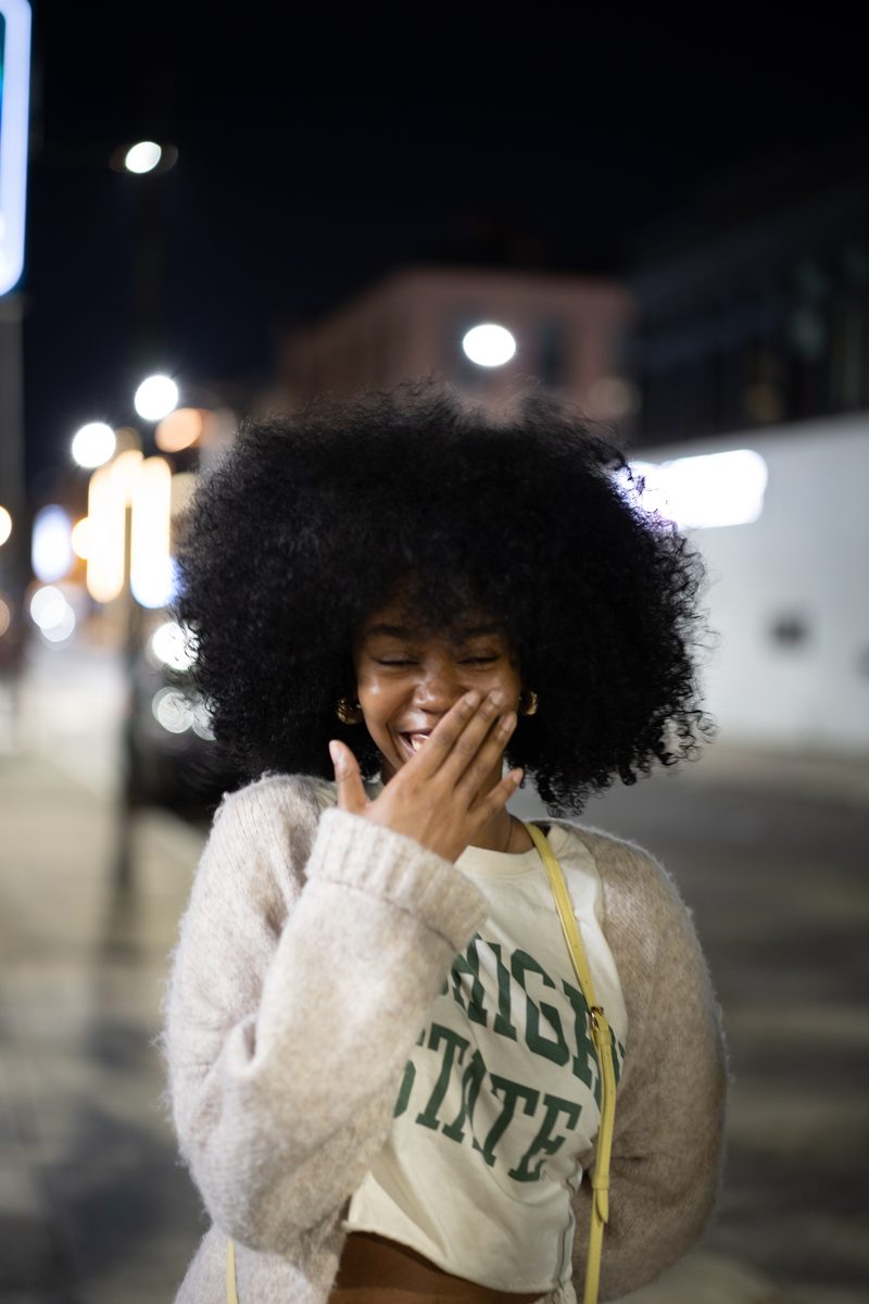 A young woman with a full afro laughs behind her hand in a Michigan State sweater on South Street at night.