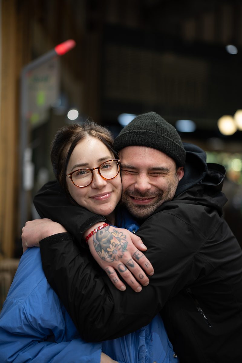 A woman in round glasses is wrapped in a hug from a man in a black beanie inside a South Street shop.