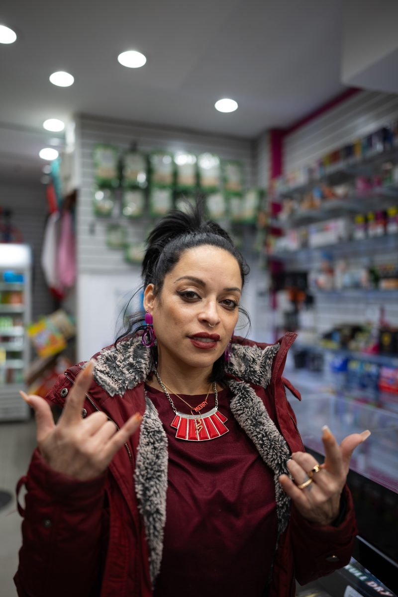 A woman in a maroon jacket with a fur collar throws up two rock-and-roll hand signs inside a South Street smoke shop.