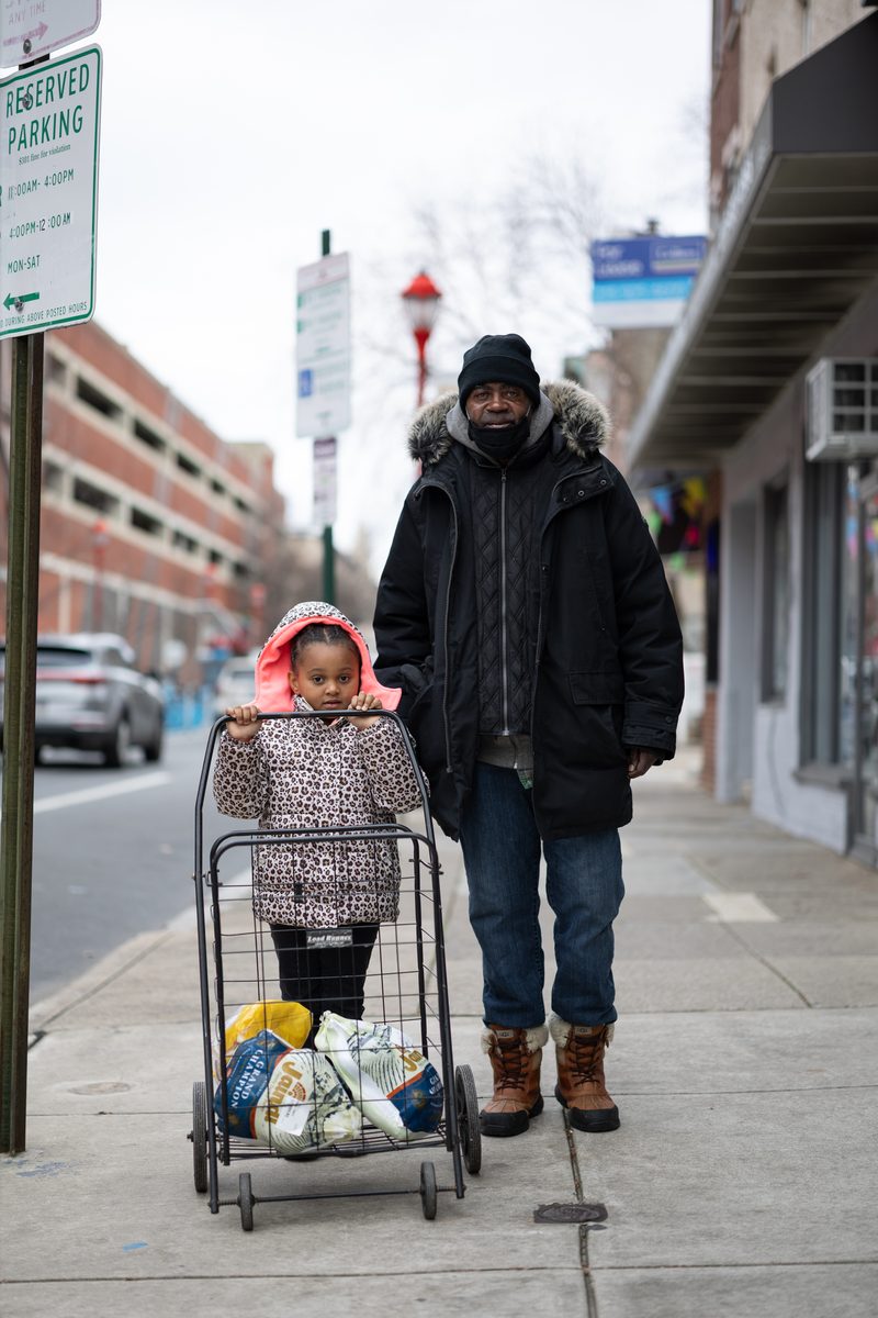 A grandfather in a winter coat and his young granddaughter pose for a photo on South Street, with the granddaughter pushing a shopping cart along the sidewalk.