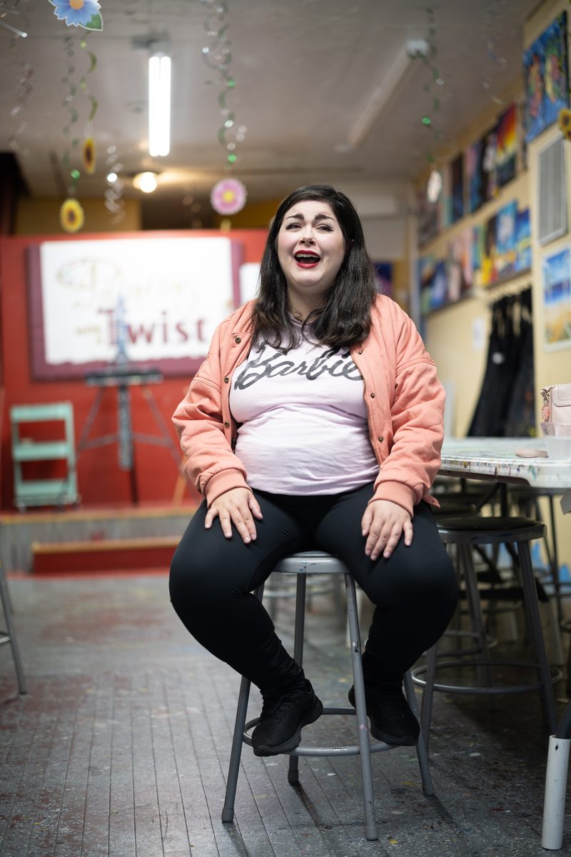 A woman in a Barbie t-shirt and pink jacket laughs, seated on a stool inside a Painting with a Twist on South Street.