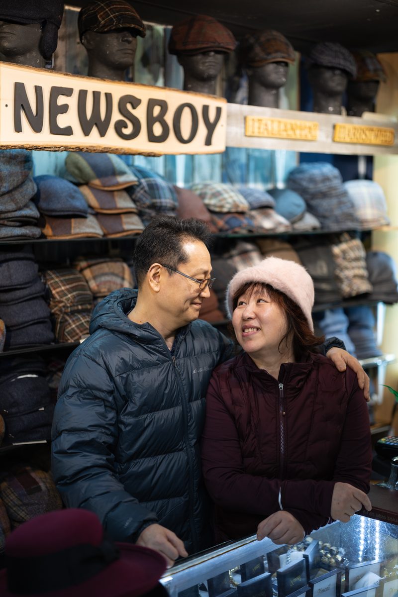 An older couple smiles at each other inside a South Street hat shop called Newsboy, with many caps stacked behind them.