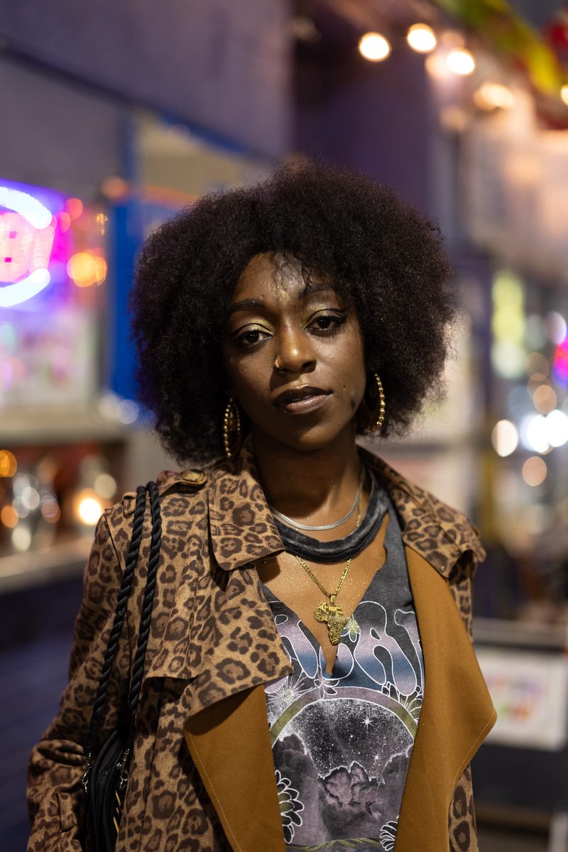 A woman with a natural afro in a leopard-print coat looks at the camera under South Street neon at night.