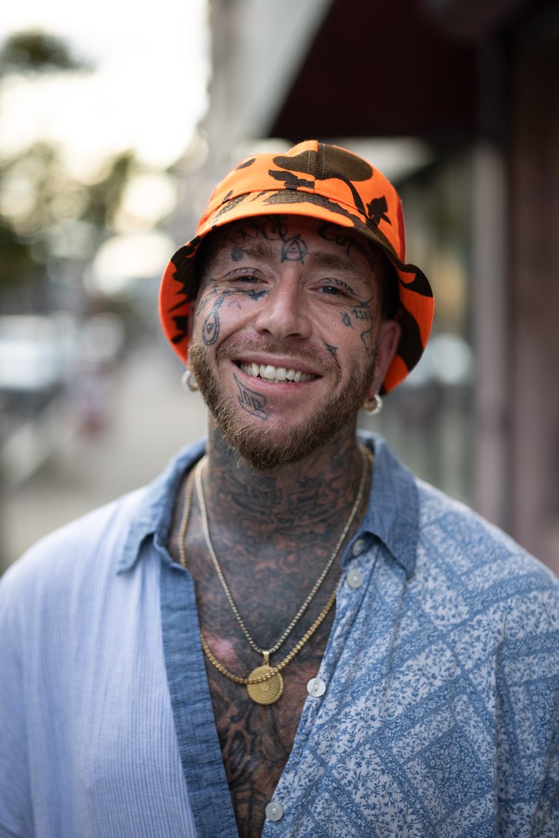A man with face tattoos in an orange camo bucket hat and blue button-up smiles on South Street.