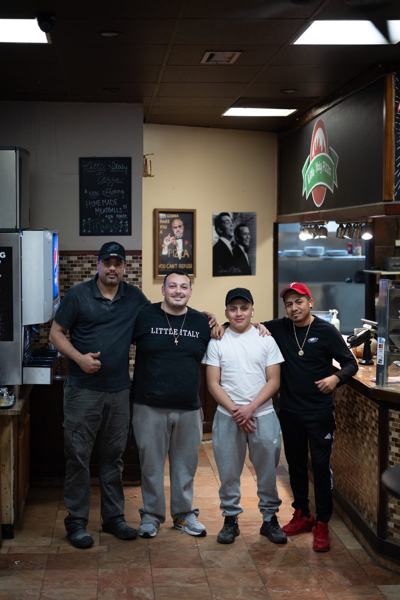 Four men stand together behind the counter of Little Italy pizza shop on South Street, one in a "Little Italy" t-shirt.