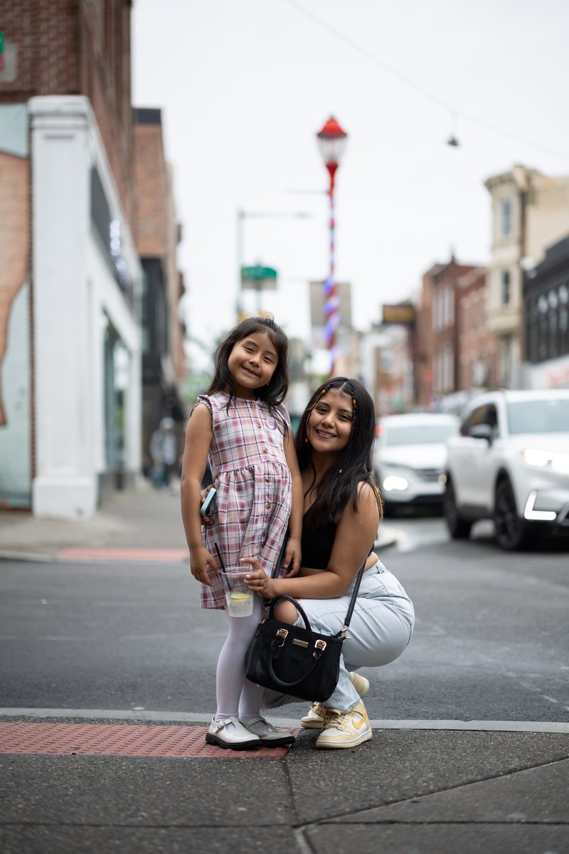 A young mother crouches beside her daughter in a plaid dress at a South Street crosswalk, both smiling.