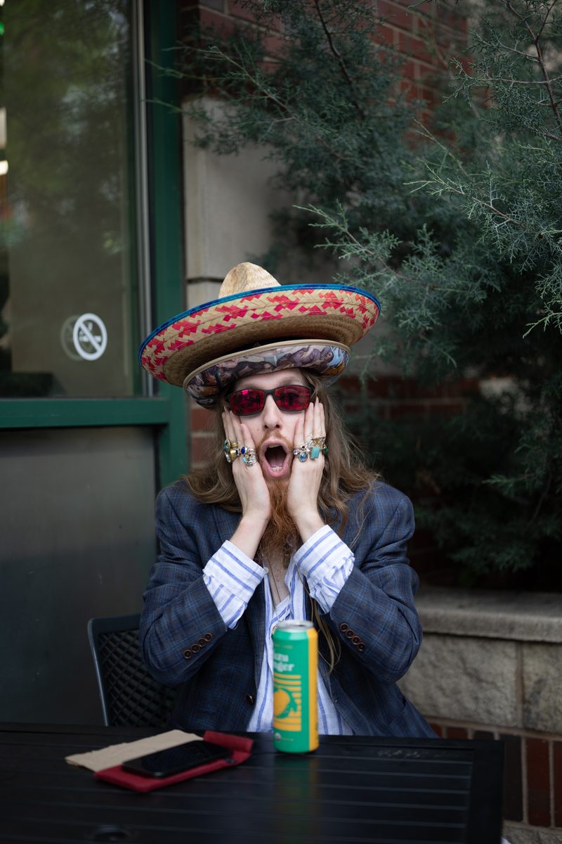 A man in a double sombrero, pink sunglasses, and a suit jacket makes a shocked face with both hands on his cheeks at outdoor café seating by Whole Foods supermarket on South Street.