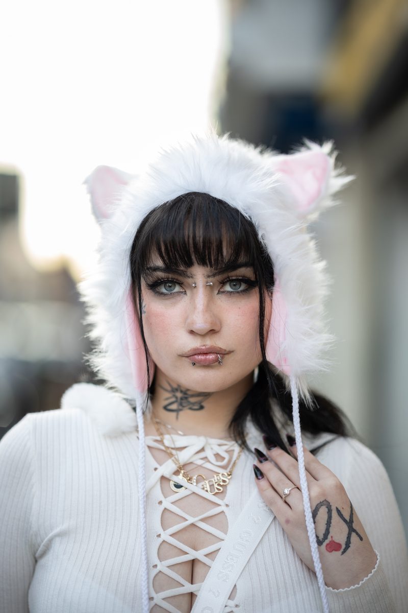 A woman in a white cat-ear hood with pink lining, lace-up top, and facial piercings looks into the camera posing for a portrait on South Street.