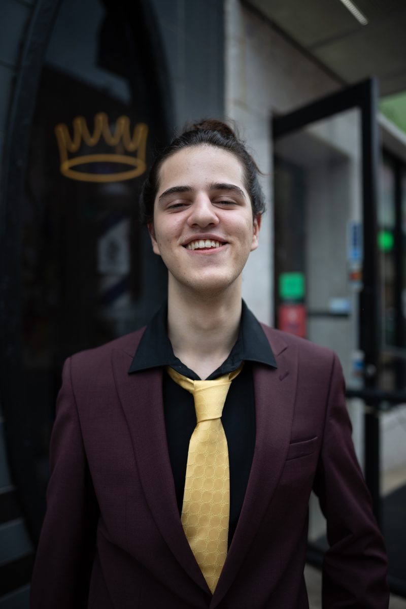 A young man in a maroon suit and gold tie smiles with his eyes closed outside a South Street shop.