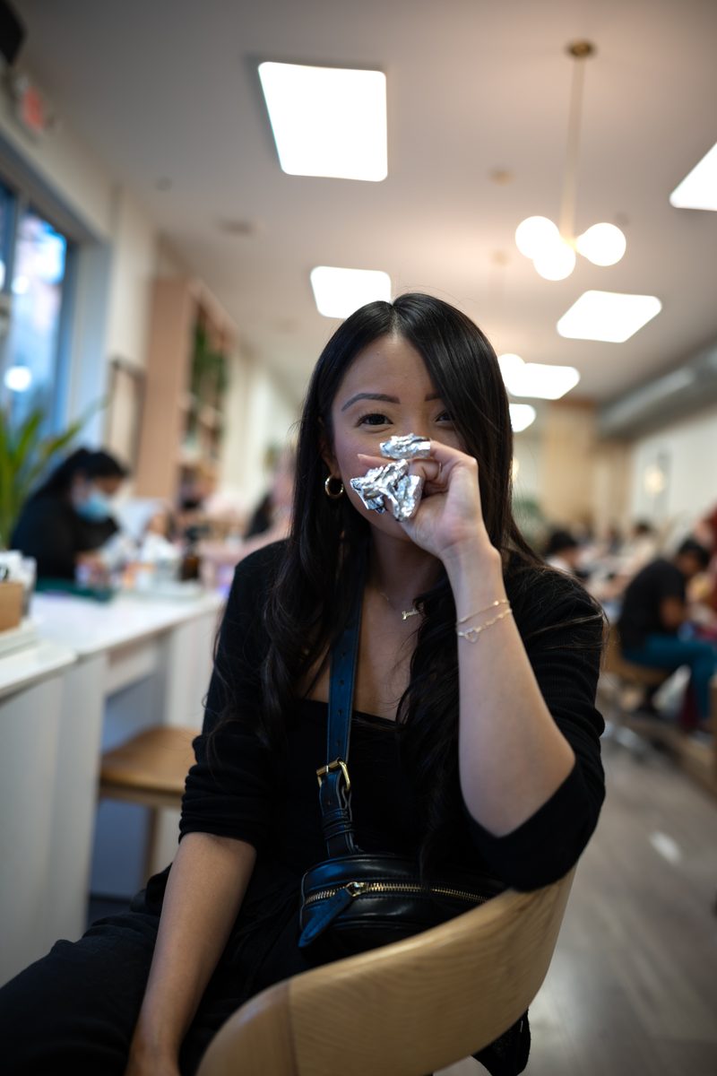 A shy, laughing young woman in black holds her foil-wrapped nails over her face, seated at Boutique Nail Cafe, a spa and nail salon on South Street.