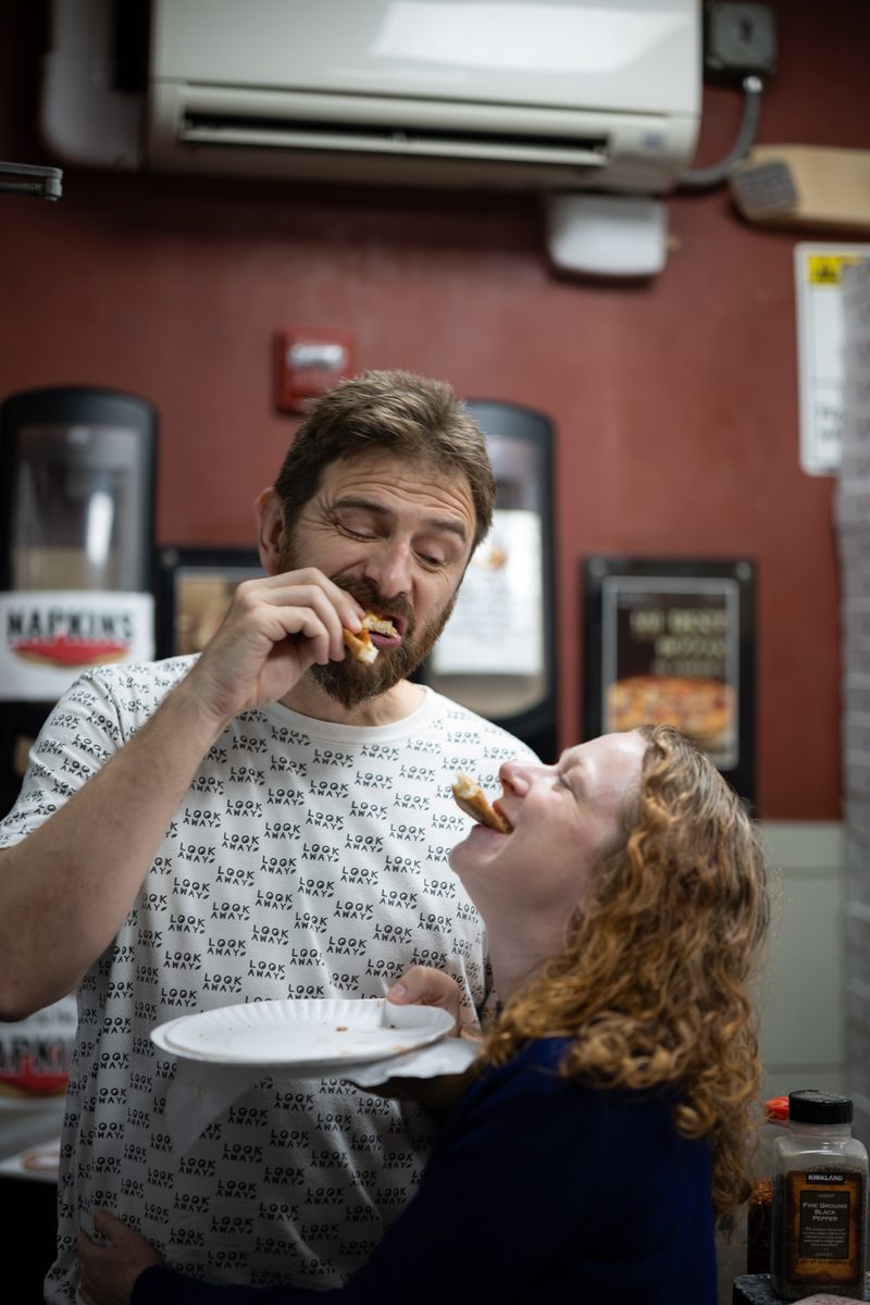 A man and laughing woman with red curls eat the crusts of pizza slices at Lorenzo and Son's Pizza on South Street.