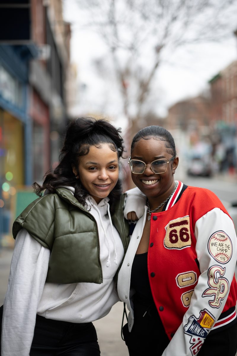 Two friends smile arm in arm on South Street, one in an olive puffer vest, the other in a red varsity jacket.