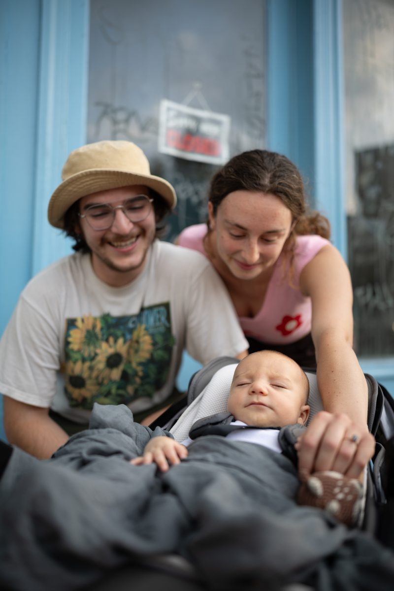 A young couple in a yellow hat and pink top holds their newborn baby, wrapped in a blanket, outside a blue South Street laundromat.