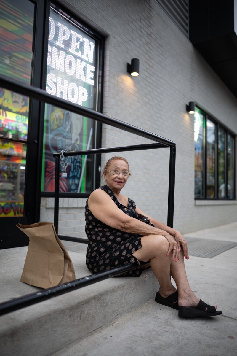 An older woman in a black floral dress sits on the steps outside a South Street smoke shop waiting for the bus, a paper bag of groceries at her side on the steps.