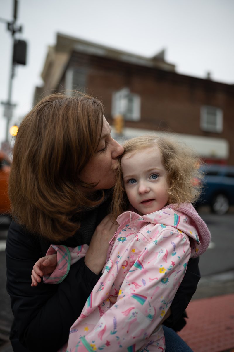 A mother kisses the cheek of her toddler daughter in a pink jacket on a South Street corner.