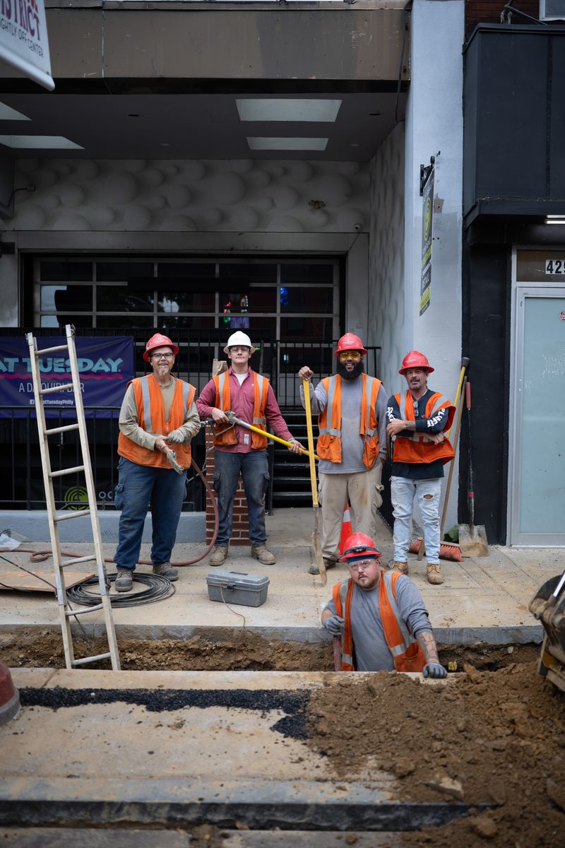 Five Philadelphia Gas Works construction workers in orange safety vests and hard hats pose at a dug-up section of South Street sidewalk, one standing in the trench.